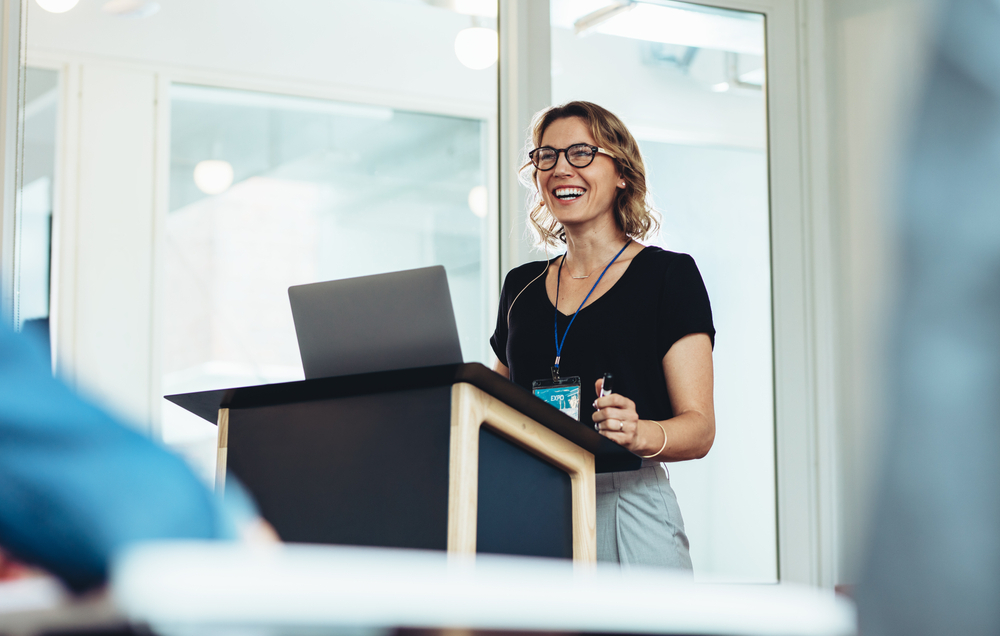 Businesswoman standing at a podium with a laptop, delivering a speech on the importance of choosing the right audio visual equipment supplier for business success.